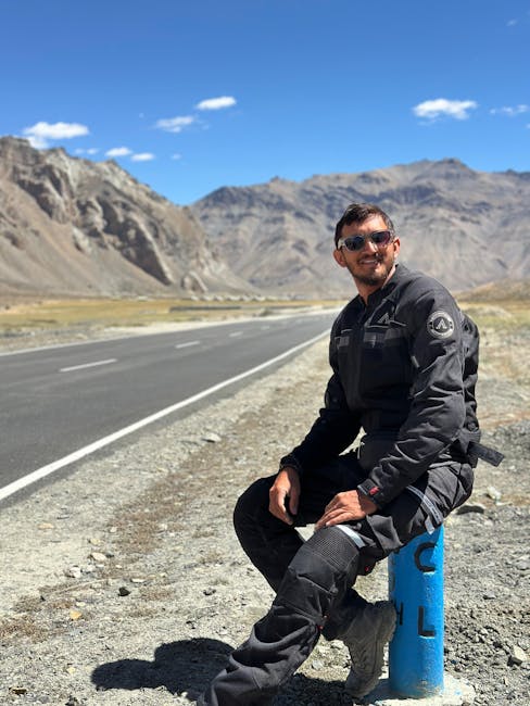 A man wearing black protective gardening and landscaping clothing, including gloves and sunglasses, sitting on a blue roadside marker post beside a paved road in a rural outdoor setting. Behind him, t